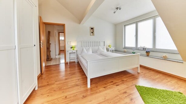 A bedroom with a white bed, wooden flooring, a green rug, a nightstand, and a window with blinds.