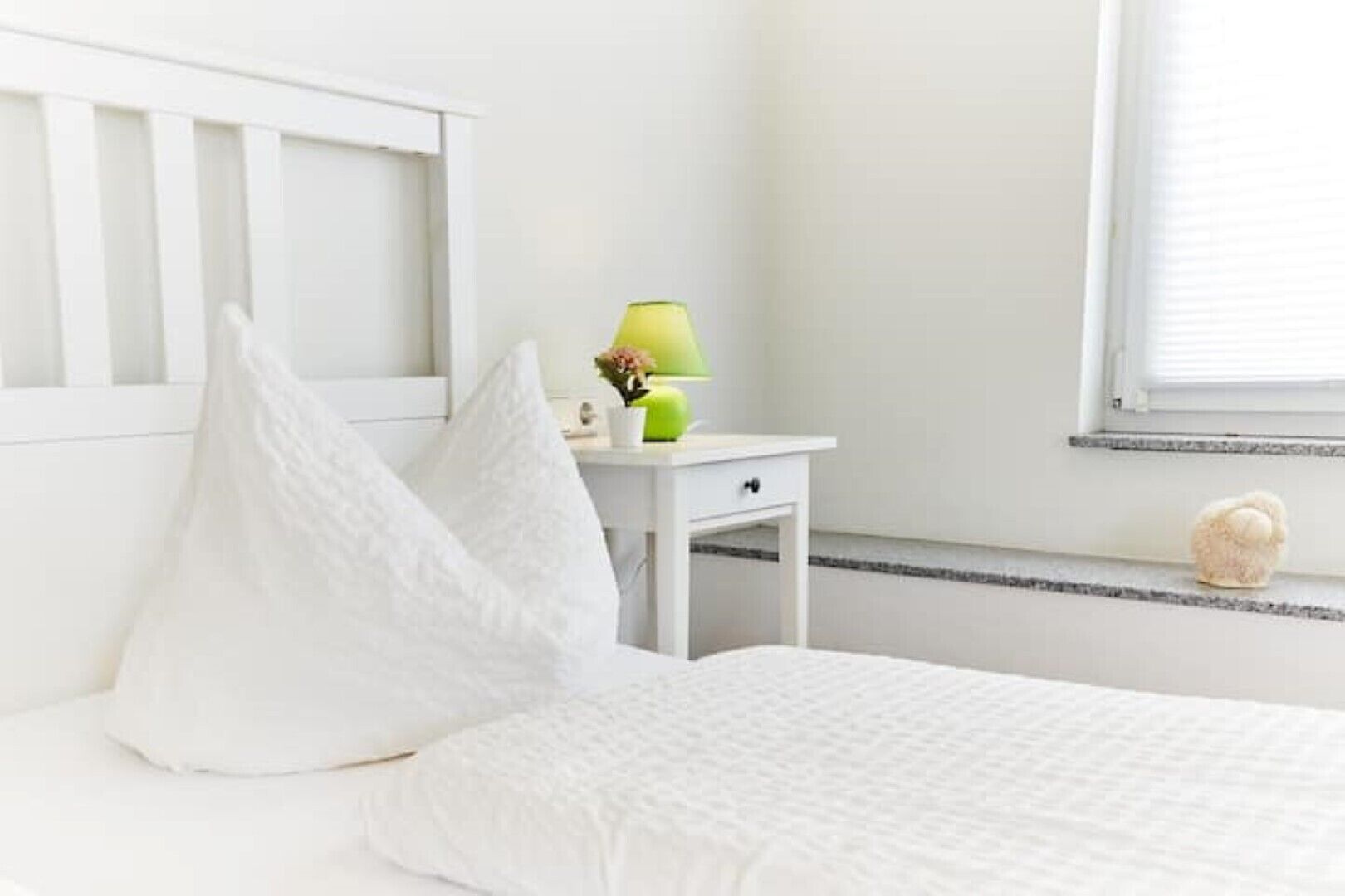 Un dormitorio blanco con cama, dos almohadas, una mesita de noche con lámpara y ventana con persianas.