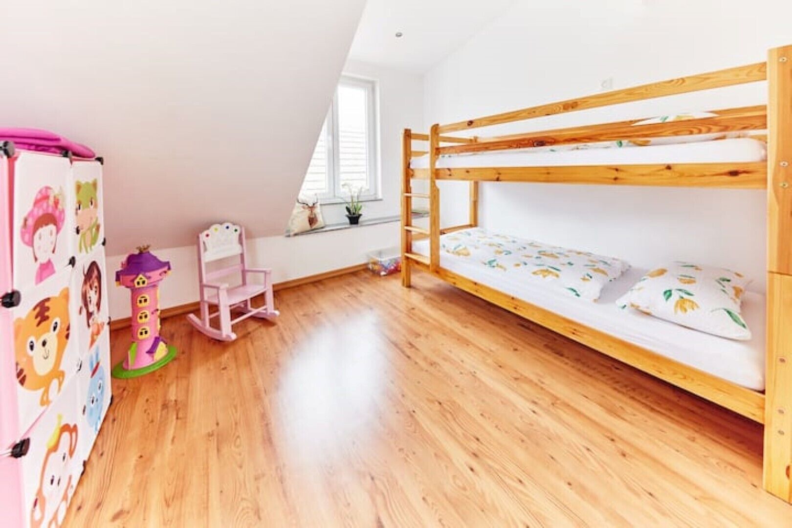 Ein Kinderzimmer mit Etagenbett, Holzboden, einem rosa Stuhl und einer Wand mit Zeichentrickfiguren.