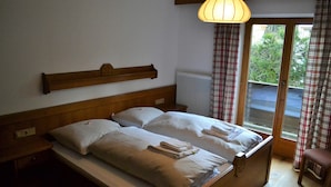 Una cama doble con dos almohadas y una toalla doblada, cabecero de madera, mesita de noche, radiador y ventana con cortinas.