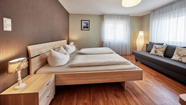 Ein modernes Schlafzimmer mit einem hölzernen Bett, einem Nachttisch mit Lampe, einem dunkelgrauen Sofa mit Zierkissen und einem großen Fenster mit transparenten Vorhängen.