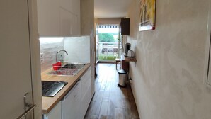 A narrow kitchen with a sink, stove, and dishwasher. A small trash can and a shelf with a kettle are visible.