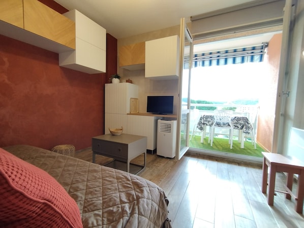 Een slaapkamer met een bed, een bureau, een televisie en een balkon met stoelen.