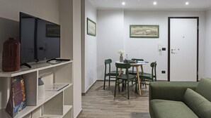 Una sala de estar moderna con un sofá verde, un televisor sobre un mueble blanco, una mesa de comedor con sillas y una puerta al fondo.