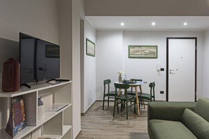 Una sala de estar moderna con un sofá verde, un televisor sobre un mueble blanco, una mesa de comedor con sillas y una puerta al fondo.