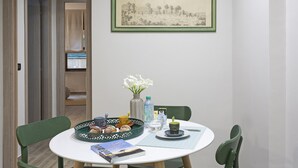 Un comedor con una mesa redonda, sillas, un cuadro en la pared, un florero con flores y una bandeja con pasteles.