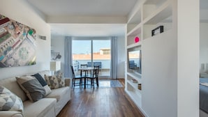 Una sala de estar moderna con un sofá, una mesa de comedor y sillas. Hay un ventanal con vistas a edificios y un balcón.