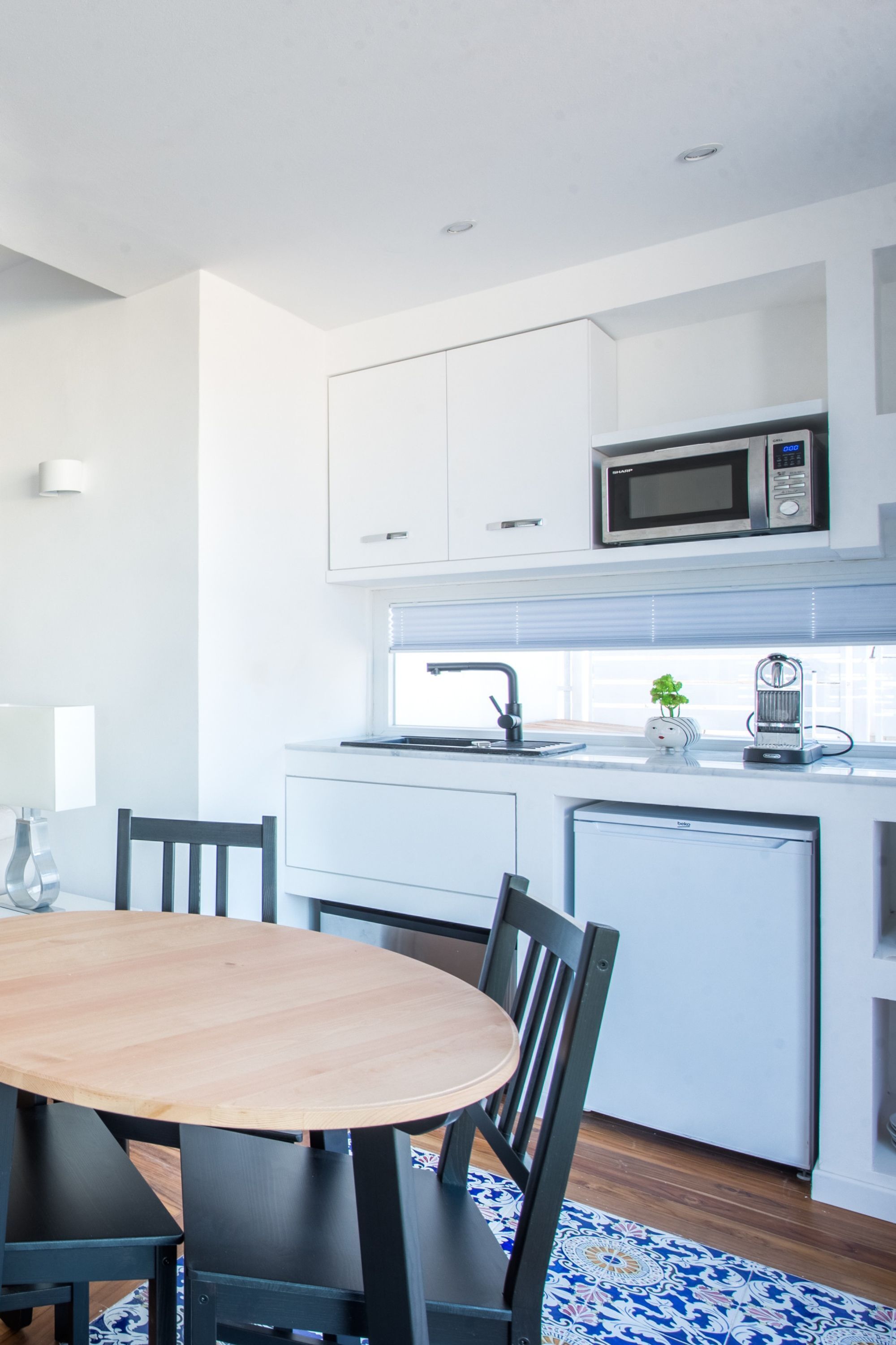 A modern kitchen with white cabinets, a microwave, and a sink. There is a dining table with chairs and a patterned rug.