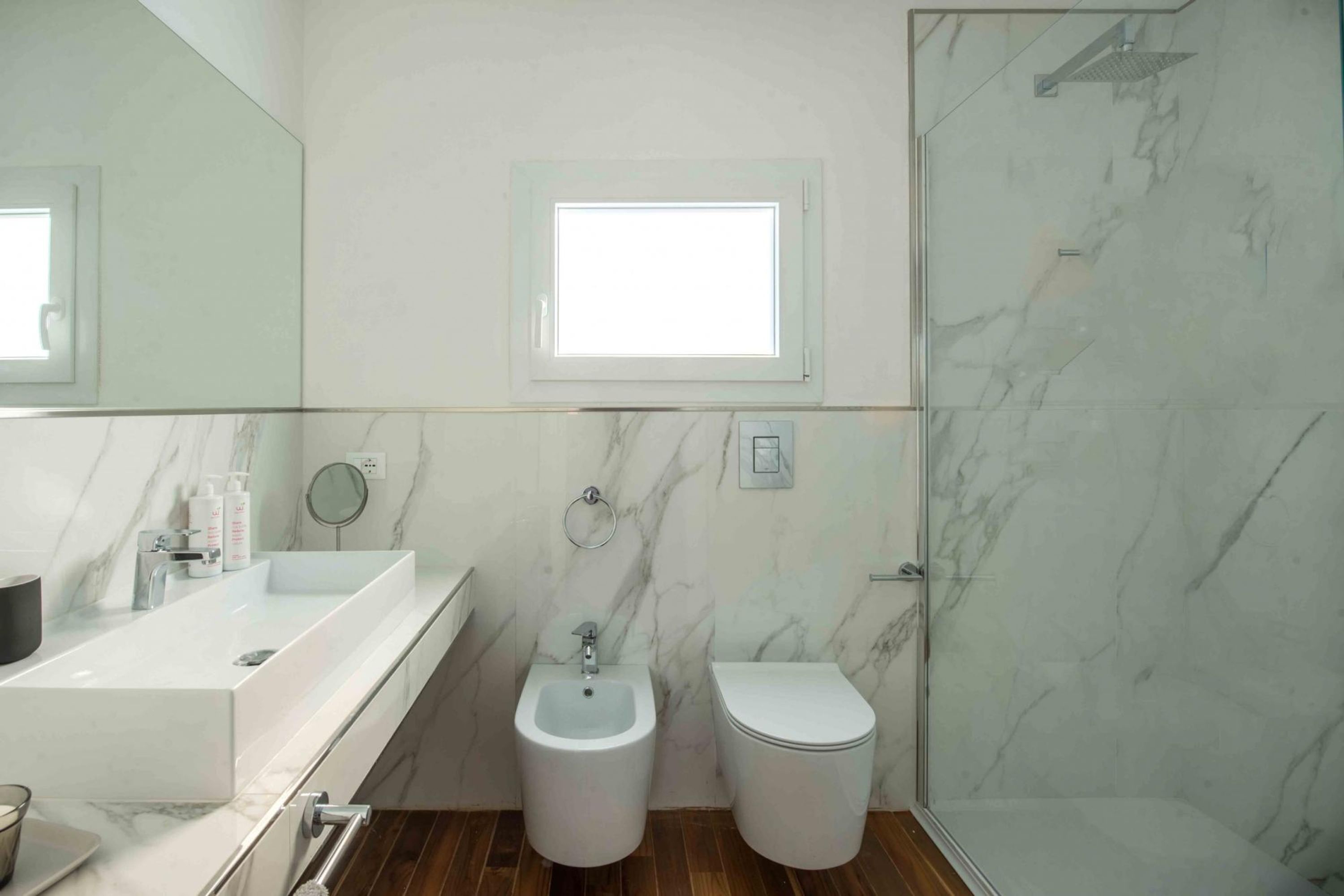 Une salle de bain moderne avec un lavabo en marbre, un bidet et des toilettes, un grand miroir et une douche dotée d’une porte en verre.