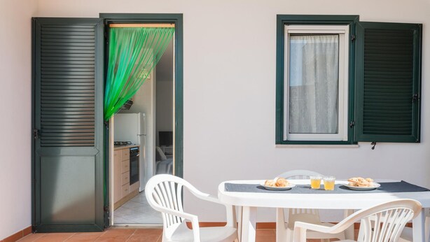 A small dining area with a table and chairs, a green curtain, and a window with shutters. - White Sea - Villetta Ariel (Carovigno)