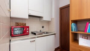 Cocina compacta con armarios blancos, un microondas rojo, un lavabo y una estantería de madera con libros.
