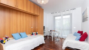 Una habitación con dos camas, un armario de madera, una mesita pequeña y una ventana con cortinas translúcidas.