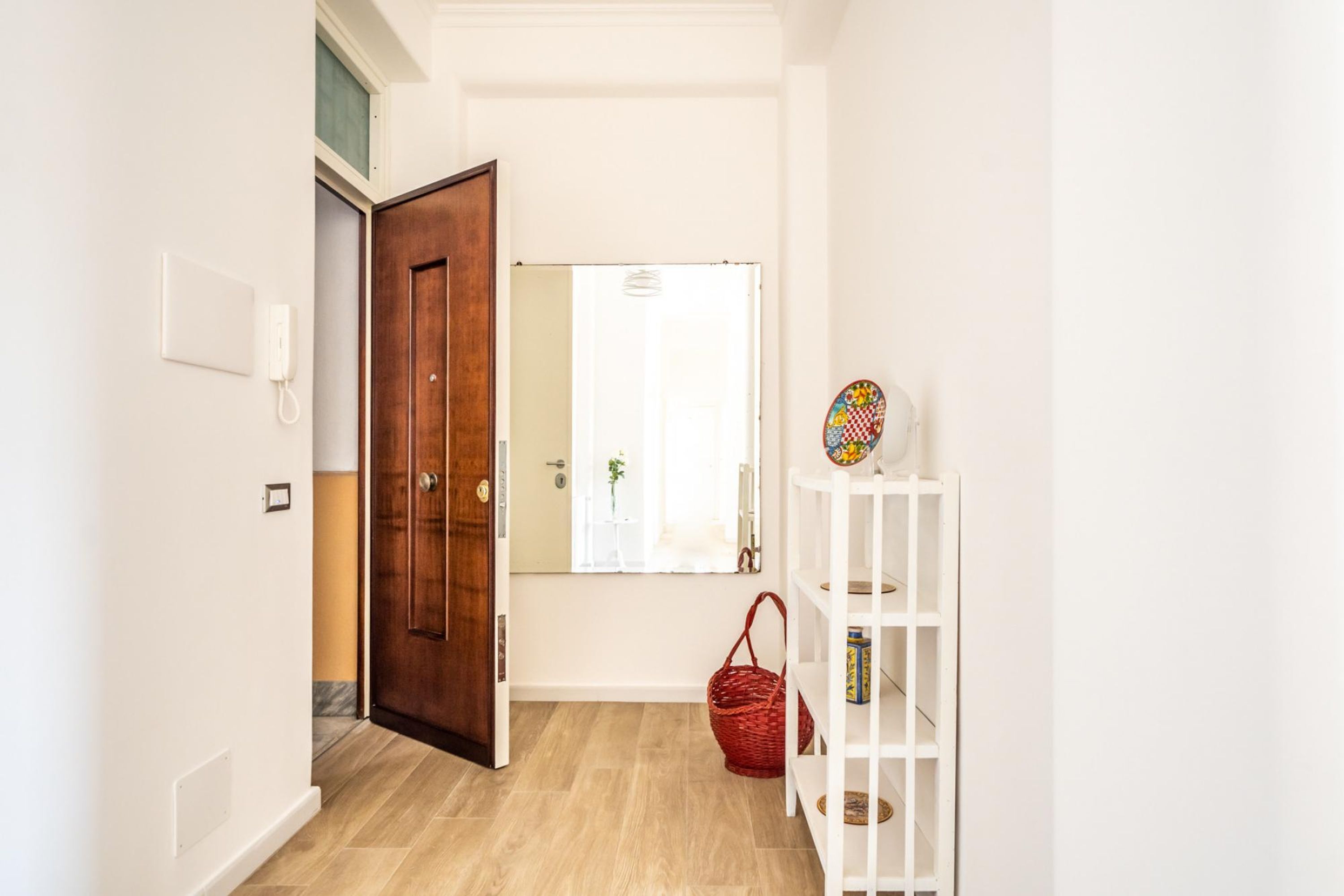 A hallway with a wooden door, a white wall-mounted phone, a mirror, a white shelf with decorative items, and a red basket.