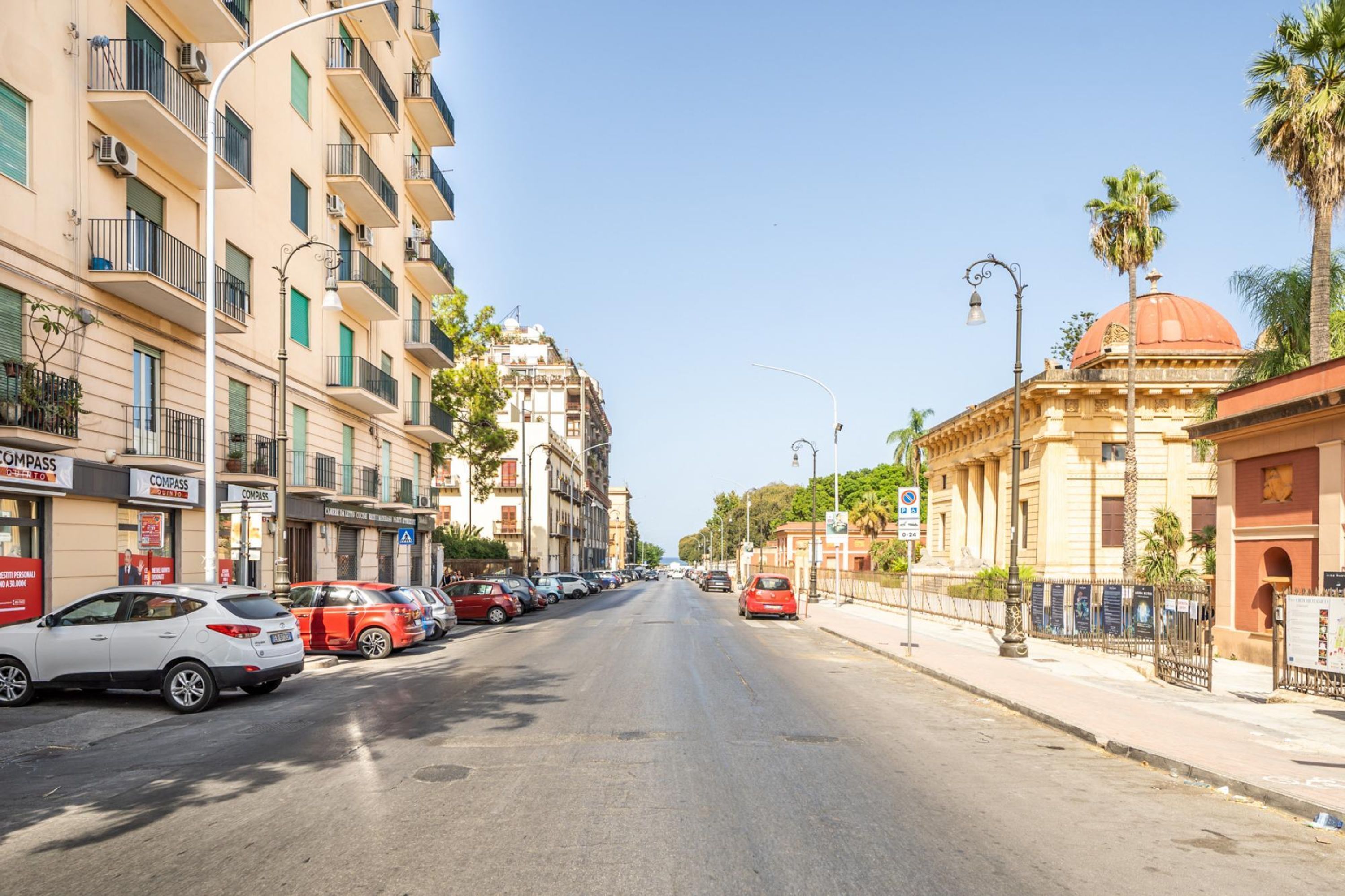 Una strada fiancheggiata da edifici a più piani, auto parcheggiate e palme.