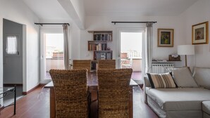 A living room with a sofa, a dining table with chairs, a bookshelf, and a window with curtains.