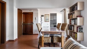 A dining area with a wooden table and chairs, a bookshelf, and a kitchen in the background.