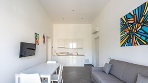 Ein modernes Wohnzimmer mit einem grauen Sofa, einem Esstisch mit weißen Stühlen, einer Küchenzeile mit weißen Schränken und einem farbenfrohen, abstrakten Gemälde an der Wand.