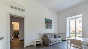 Ein modernes Wohnzimmer mit einem grauen Sofa, einem kleinen weißen Tisch und einem Esstisch mit Stühlen. An der Wand hängt ein farbenfrohes abstraktes Gemälde und ein Fenster bietet Blick auf Gebäude.