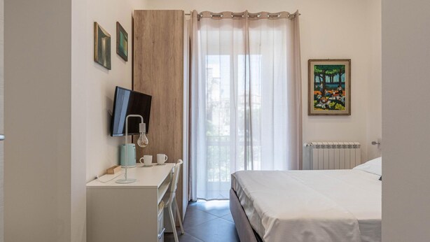 Ein Schlafzimmer mit einem Bett, einem Schreibtisch mit Computer, einem Stuhl und einem Fenster mit Vorhängen.
