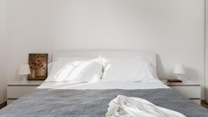 A neatly made bed with white linens and a gray quilt, two bedside lamps, and a painting on the nightstand.