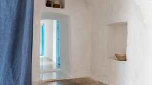 A narrow hallway with white walls, a tiled floor, and a blue curtain on the left. There are two doors, one leading to a brighter area and another to a room with a small shelf holding a bowl.