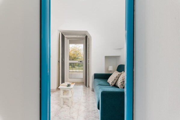 A narrow hallway with a door leading to a balcony, a teal sofa with patterned cushions, and a small white table with a book and a cup.