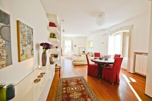 Una sala de estar moderna con zona de comedor, que presenta piso de madera, sillas rojas, alfombra estampada y sofá blanco.