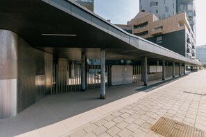 Eine moderne Fußgängerbrücke mit flachem Dach, gestützt von Säulen, die zu einem Gebäude mit ebener Fassade und mehreren Balkonen führt.
