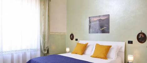 A bedroom with a bed, two bedside lamps, a nightstand, a window with curtains, and a wall-mounted picture.