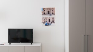 A modern room with a flat-screen TV on a white cabinet, a wardrobe, and a wall with two photos of a historic building and a blue tuk-tuk. - Dioniso Smart Suite With Balcony by Wonderful Italy (Syracuse)
