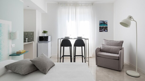 A modern, minimalist living room with a sofa, two chairs, a dining table with chairs, a small kitchen area, and a floor lamp. - Dioniso Smart Suite With Balcony by Wonderful Italy (Syracuse)