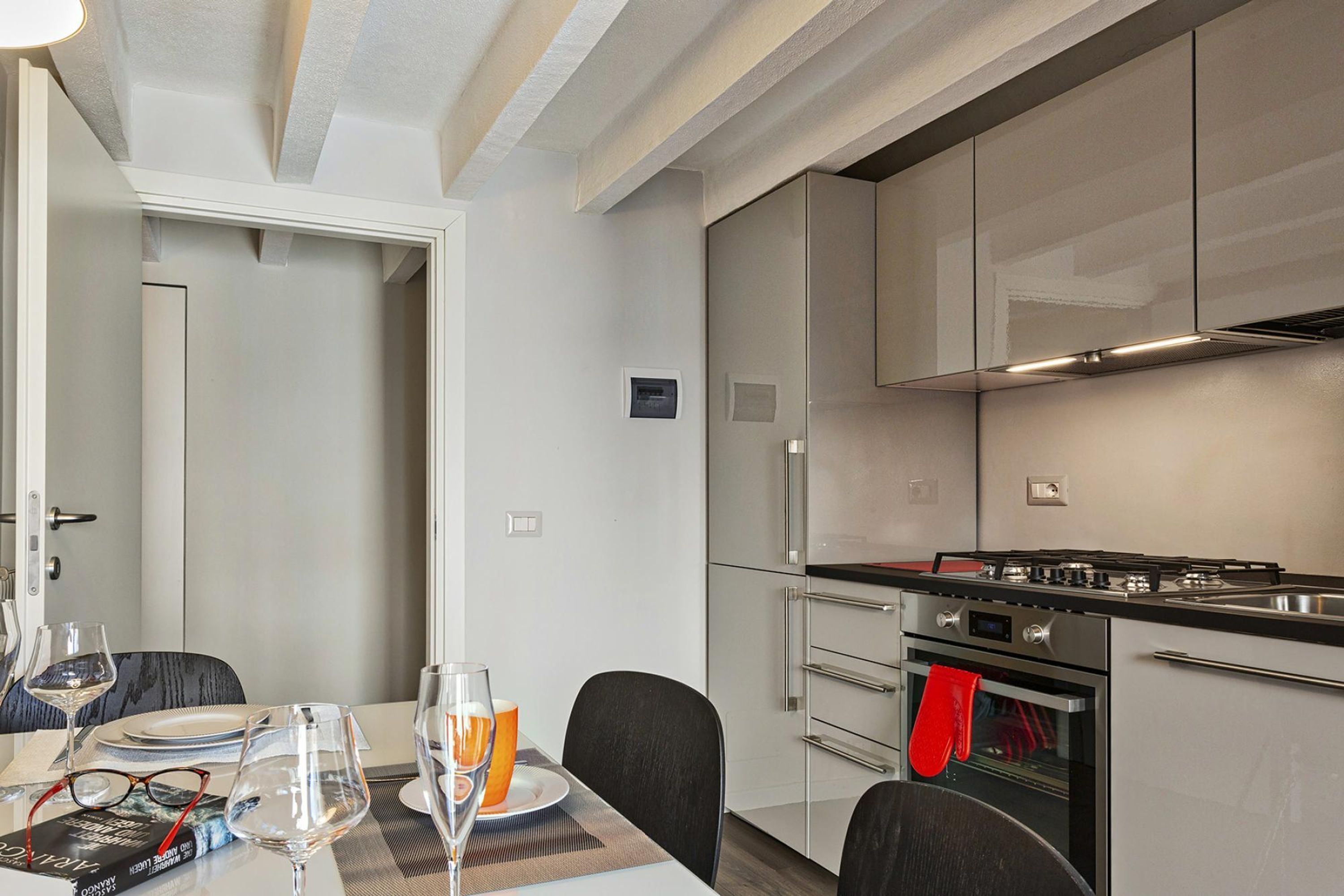 A modern kitchen with stainless steel appliances, a dining table set with glasses and plates, and a red oven mitt hanging on the oven handle.