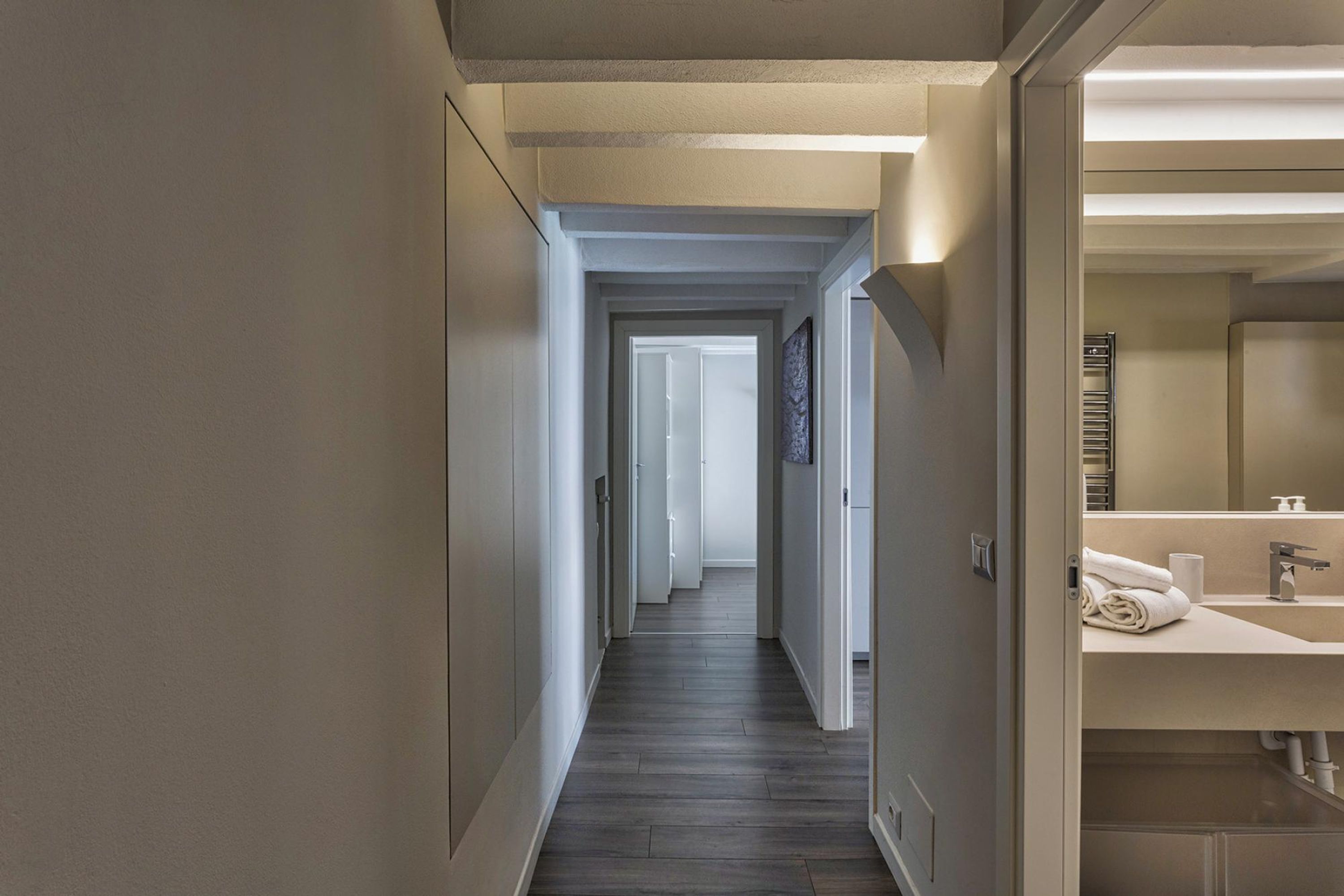 A hotel hallway with a wooden floor, a bathroom with a sink and towel rack, and a door leading to another room.