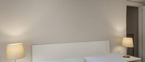 A neatly made bed with white linens in a hotel room. There are two bedside tables, each with a lamp. The walls are painted in a light color, and there is a door visible in the background.
