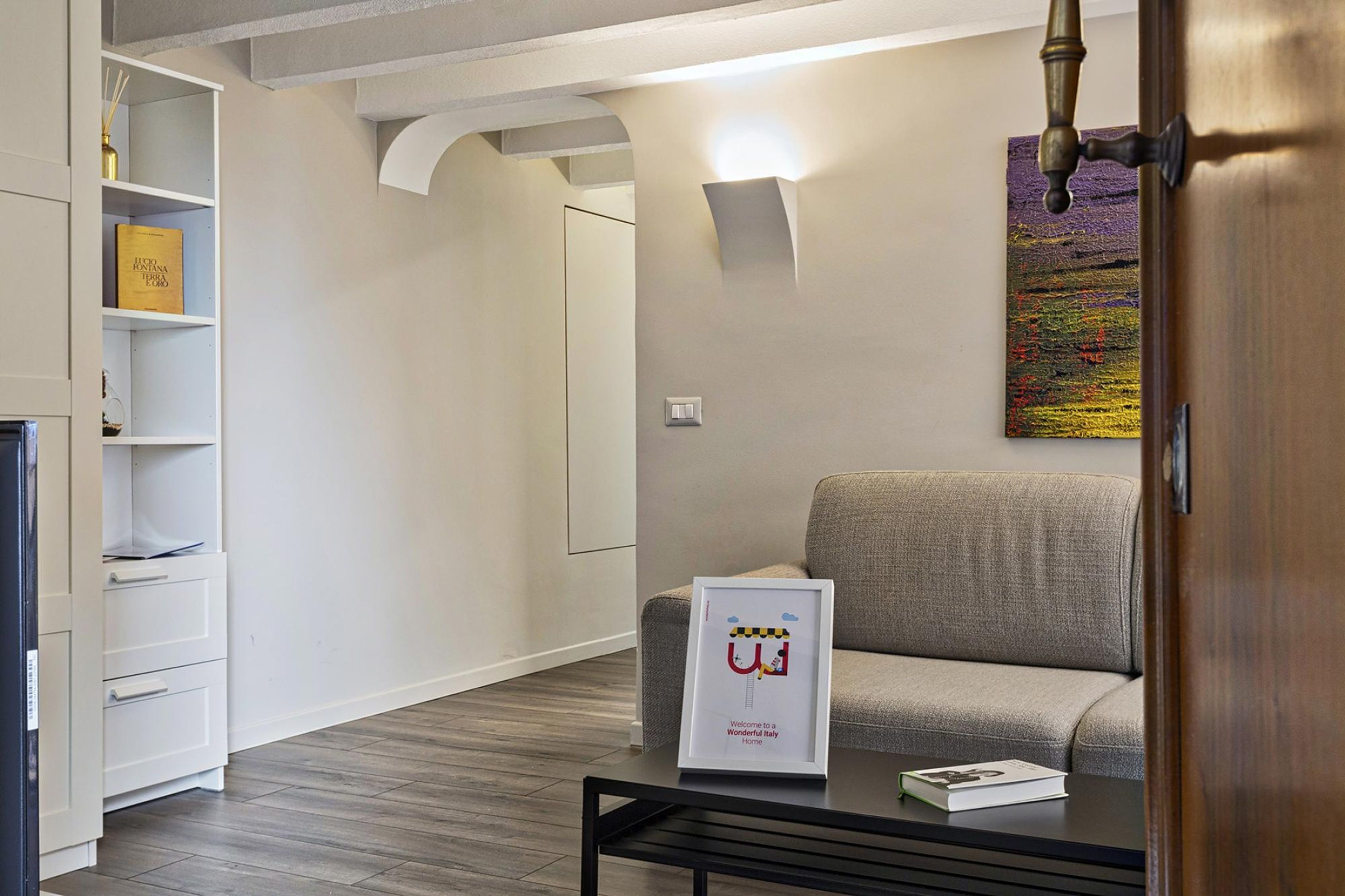 A modern living room with a sofa, a coffee table with a book, a framed welcome sign, and a bookshelf.