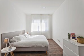 Een minimalistische slaapkamer met een bed, nachtkastje, lamp en een dressoir met boeken en een plant.