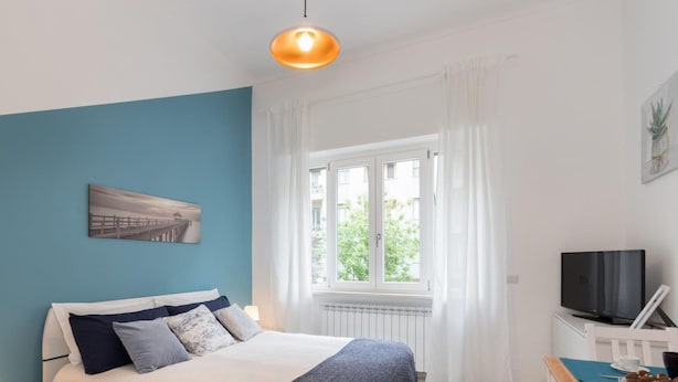 A bedroom with a bed, a window with curtains, a television, and a painting on the wall. - Studio Glicine by Wonderful Italy (Turin)