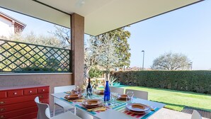 An outdoor dining area with a table set for four, a red cabinet, and a view of a garden and hedge.