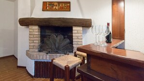 A cozy room with a stone fireplace, wooden mantel, and a painting above it. There is a table with wine glasses and a bottle.
