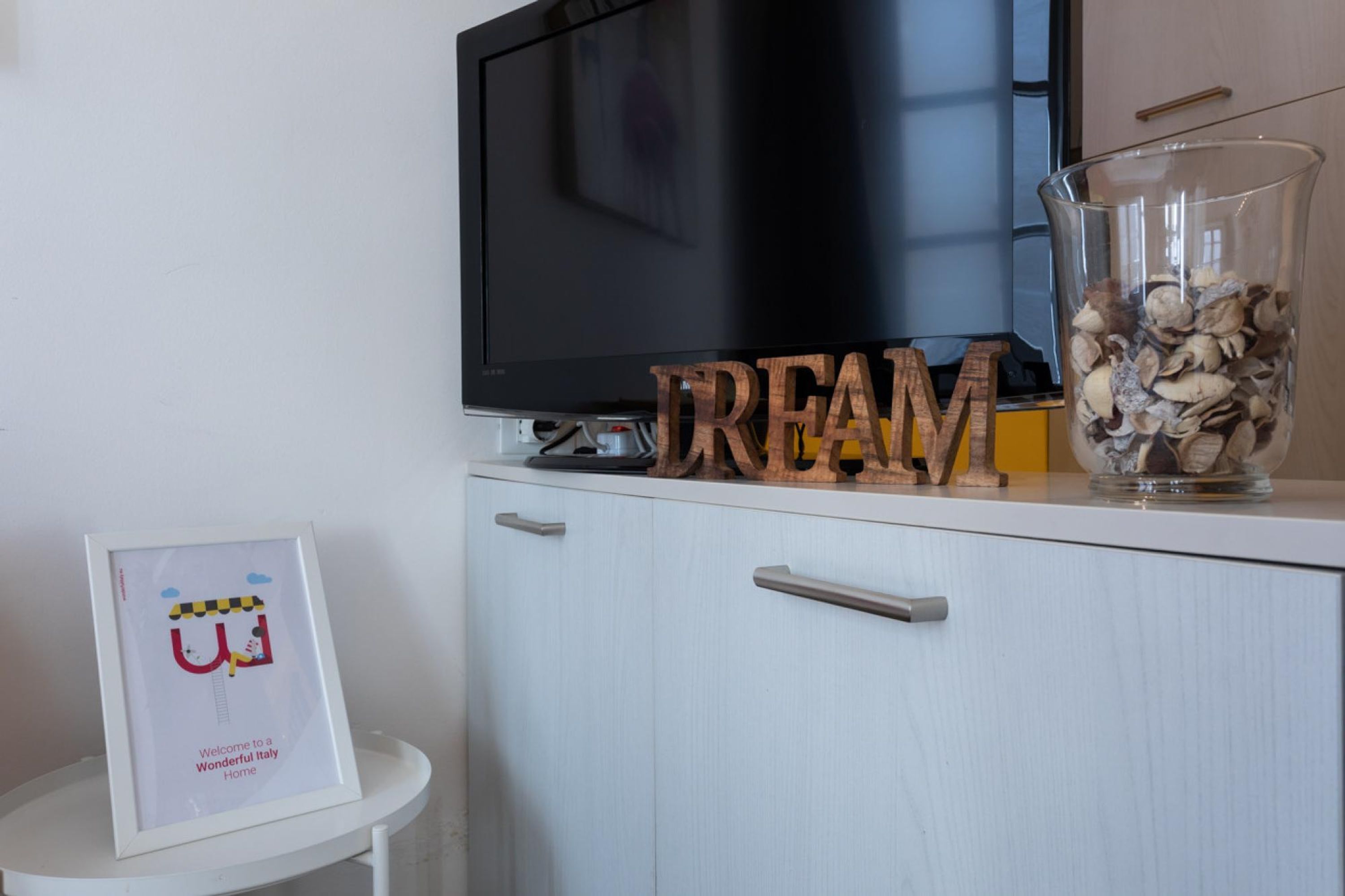 Una sala moderna con un televisor de pantalla plana, un cartel de bienvenida enmarcado, letras de madera que deletrean 'DREAM' y un jarrón de vidrio con adornos sobre un aparador blanco.