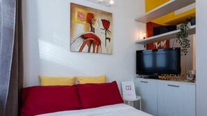 Quarto de hotel com uma cama, almofadas vermelhas e amarelas, um quadro na parede, uma televisão e uma pequena mesa com um aviso.