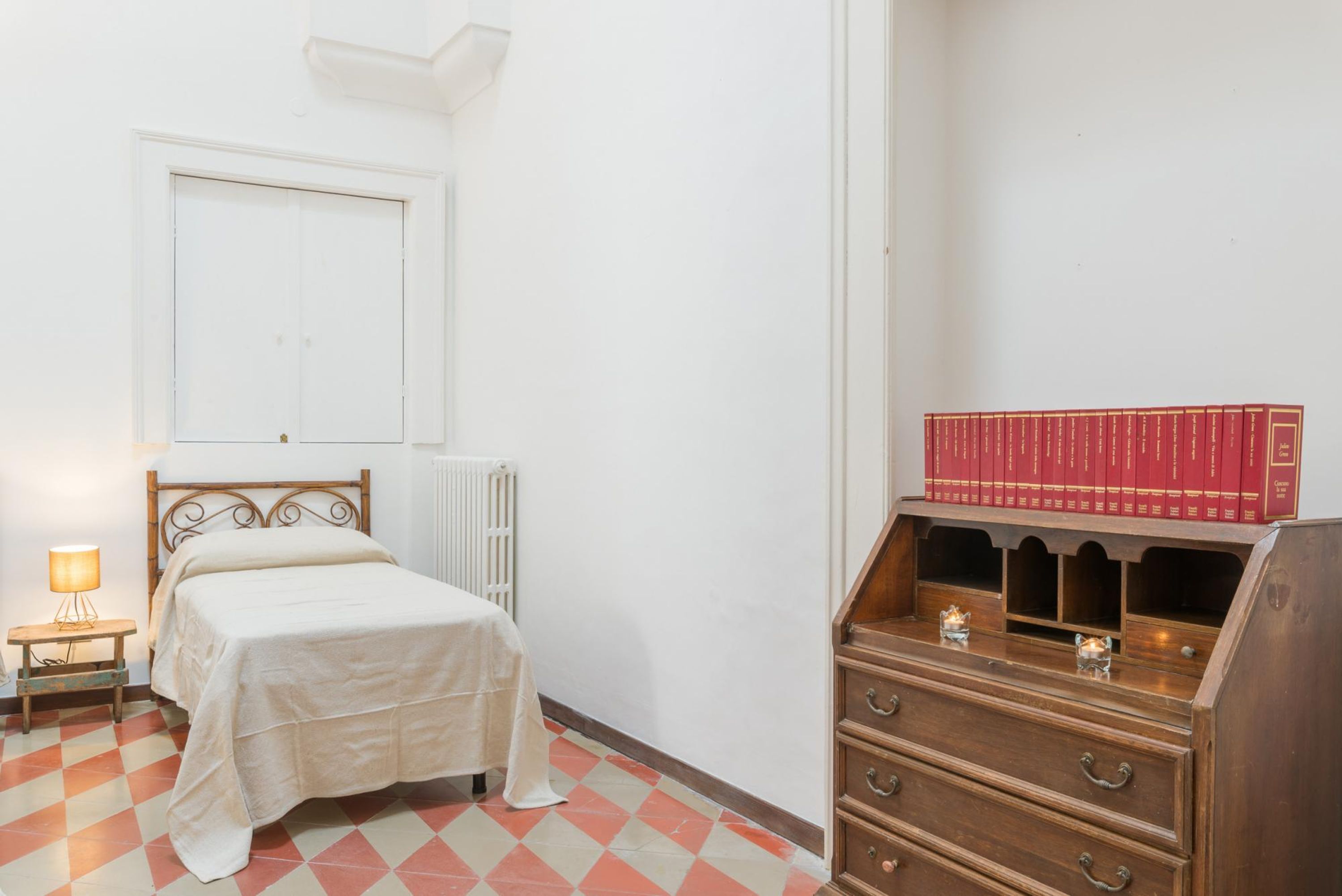 Ein Schlafzimmer mit einem Bett, einem Nachttisch mit Lampe, einer Heizung, einer Holzkommode mit Büchern und einem karierten Boden.