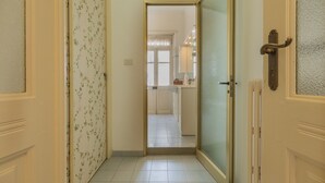 Un couloir mène à une cuisine. Une porte se trouve sur la droite et un mur recouvert d’un papier peint à motifs floraux se trouve sur la gauche.