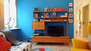 Un salon avec un mur bleu, un meuble de rangement en bois contenant des livres et un téléviseur, un fauteuil jaune et un tapis à motifs.