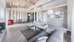 A spacious living room with a large grey sectional sofa, a glass coffee table, and a dining area with a red chair and white table.