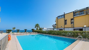 A clear blue pool with lounge chairs and umbrellas, a lifebuoy, and a view of the sea.