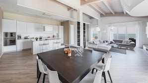 A spacious living area with a large dining table, white chairs, a sofa, and a view of the sea.