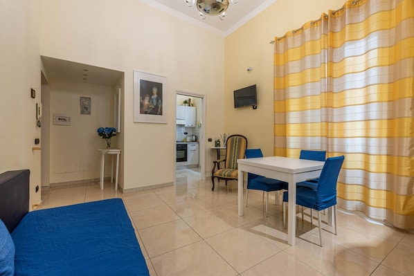 A hotel room with a bed, a dining table with chairs, a TV, and a kitchen area visible through an open door. - Olivella Apartment al Teatro Massimo by Wonderful Italy (Palermo)