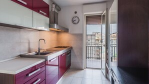 A compact kitchen with a gas stove, sink, and modern cabinetry. A clock is visible on the wall, and the kitchen leads to a balcony with a view of buildings.