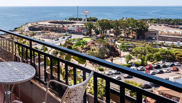 A balcony with a view of a coastal town, including a round building and parked cars.